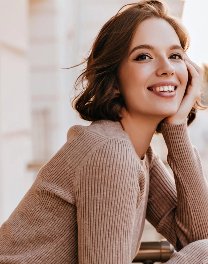 A young woman with shoulder-length, wavy brown hair smiles warmly at the camera. Her light skin glows in natural daylight, and her hand rests gently on her face. She wears a soft, beige ribbed knit sweater with long sleeves. The blurred, light-toned architectural background adds depth, focusing on her joyful expression. - Alumier MD Peels in Toronto, Ontario