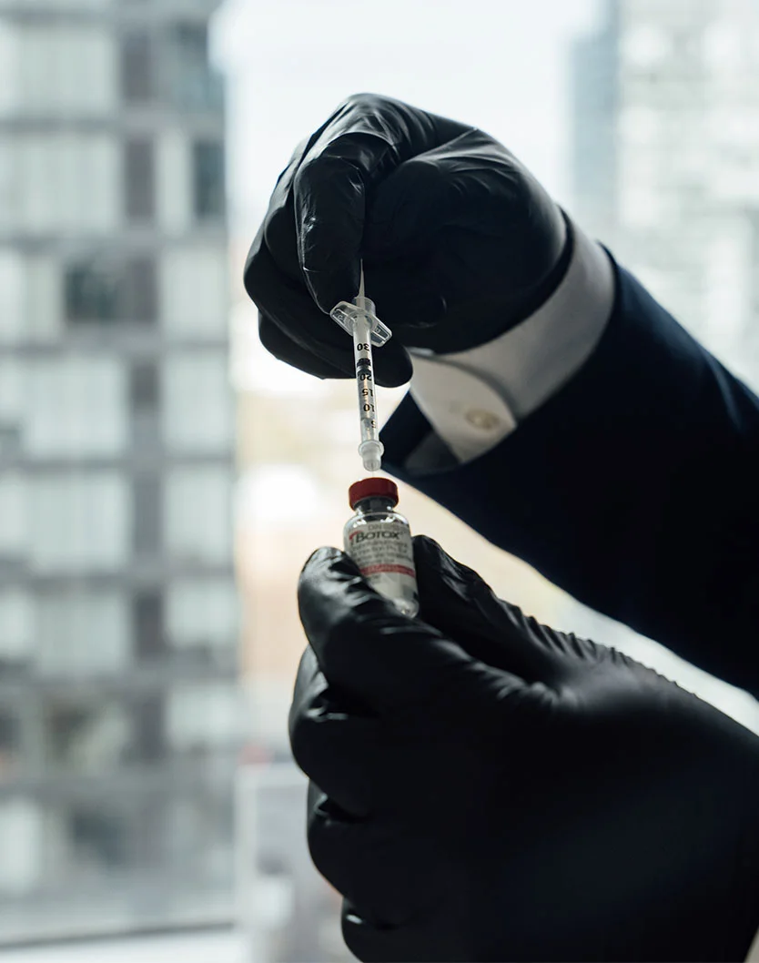 Close-up of hands in black medical gloves preparing a vaccine. One hand holds a small vial, while the other draws the liquid into a syringe. The background is blurred, showing a window with a building view. The lighting is bright and neutral, focusing attention on the hands and instruments. - Botox Dysport in Toronto, Ontario