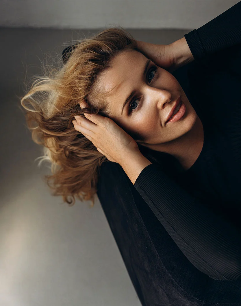 A high-angle close-up of a woman with shoulder-length, light brown/blonde hair. She holds her hair behind her head, wearing a black, form-fitting top with long sleeves. Her expression is relaxed and contemplative, with her gaze directed upward. The background is a neutral, light gray/beige wall. Soft lighting highlights her smooth, light-toned skin and subtle facial features. The overall feel is a portrait or fashion-focused shot. - Brow Lift in Toronto, Ontario