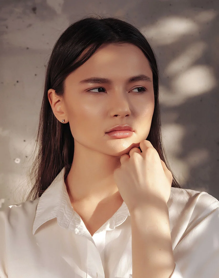 Close-up of a young woman with long, dark brown hair, gazing off to her right with a thoughtful expression. She wears a white button-down shirt, and her hand rests gently on her chin. Sunlight casts soft shadows on her face and the light-gray concrete wall behind her, with hints of leaves or branches creating patterns. - Clear and Brilliant Laser in Toronto, Ontario