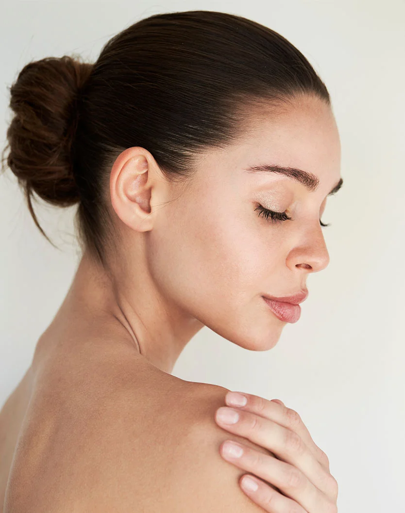Close-up of a woman with a serene expression. Her dark brown hair is in a smooth, high bun, and her eyes are closed, her face relaxed. Her light-toned skin appears smooth and healthy. She gently places a hand on her shoulder, showcasing light, well-formed nails. The background is a plain, soft beige or off-white, with even, soft lighting highlighting her features. - Earlobe Filler in Toronto, Ontario
