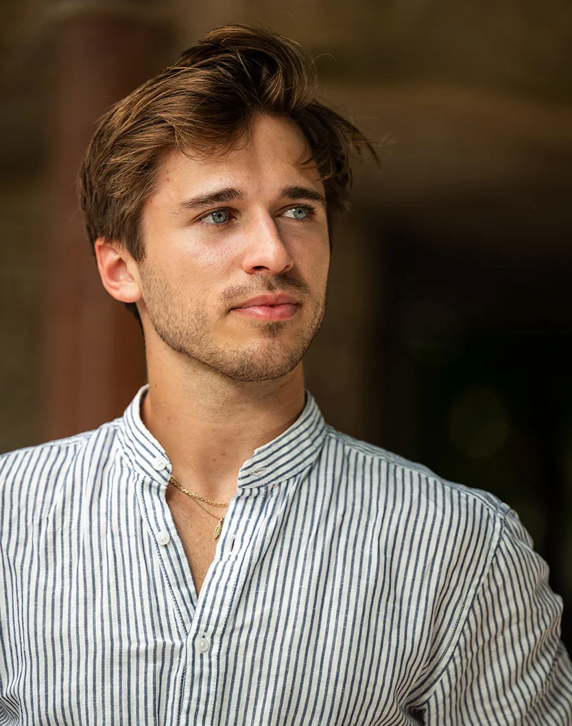 Close-up portrait of a young man with light skin and short, tousled brown hair. He has light blue eyes and a pleasant, focused expression, looking slightly to his right. He wears a light-colored, striped linen shirt with a mandarin collar and a thin gold necklace. The background is a dark, warm-toned blur, with soft, even lighting highlighting his features. - Earlobe Repair in Toronto, Ontario