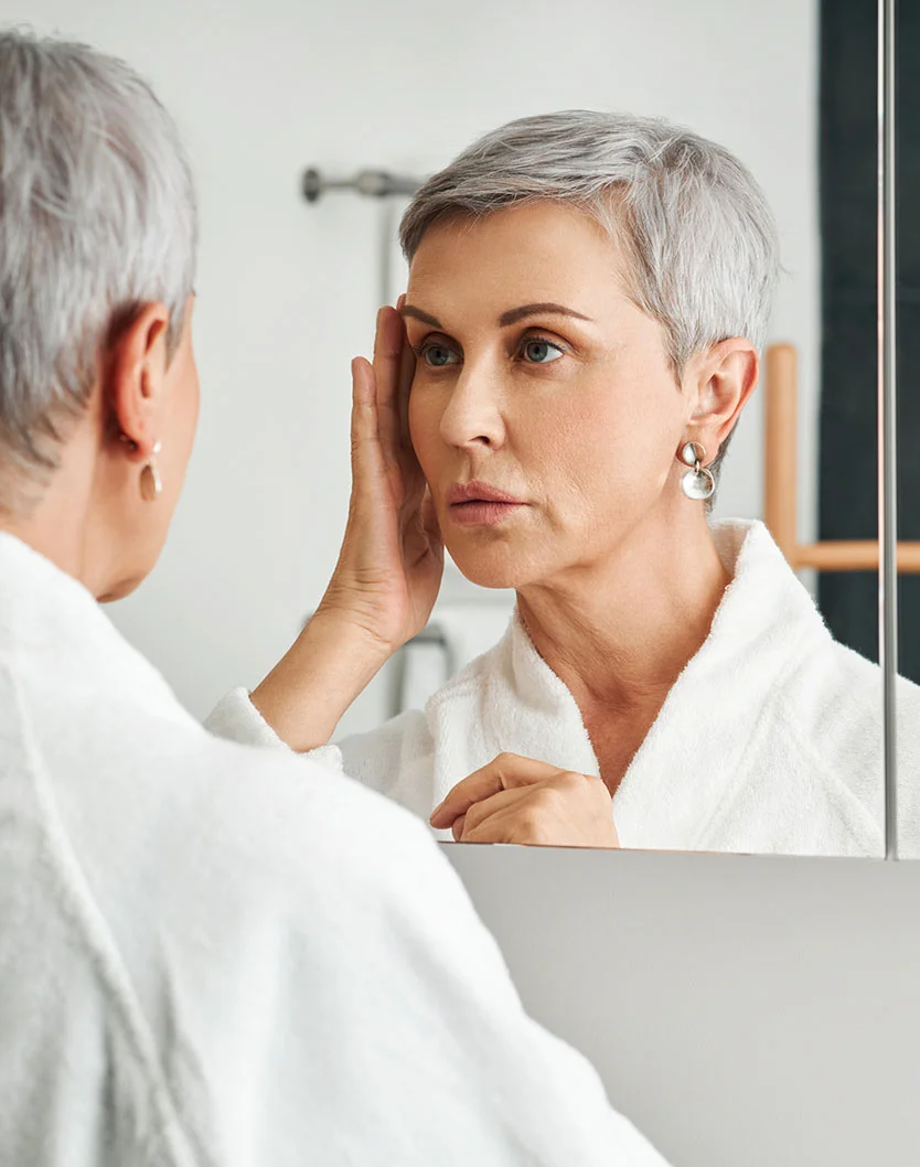 A woman with short gray hair looks at herself in a bathroom mirror, wearing a white bathrobe. Her expression appears thoughtful or concerned as she touches her forehead. The bathroom lighting is bright and neutral, and the background is light-colored, possibly part of the vanity. The focus is on her face and reflection, with the surroundings slightly blurred. - Eyelid Surgery in Toronto, Ontario