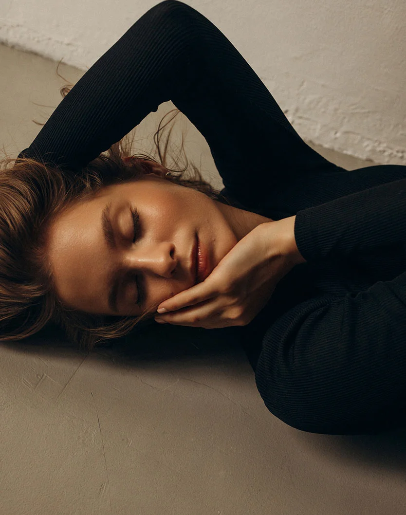 A woman lies on a textured taupe floor, wearing a black ribbed long-sleeve top. Her light brown hair is tousled around her shoulders, eyes closed, with a serene expression. One hand rests on her cheek, the other along her jawline. Soft, diffused lighting casts gentle shadows, enhancing the contours of her face. The muted cream wall in the background adds to the calm, intimate aesthetic. - Facial Slimming in Toronto, Ontario
