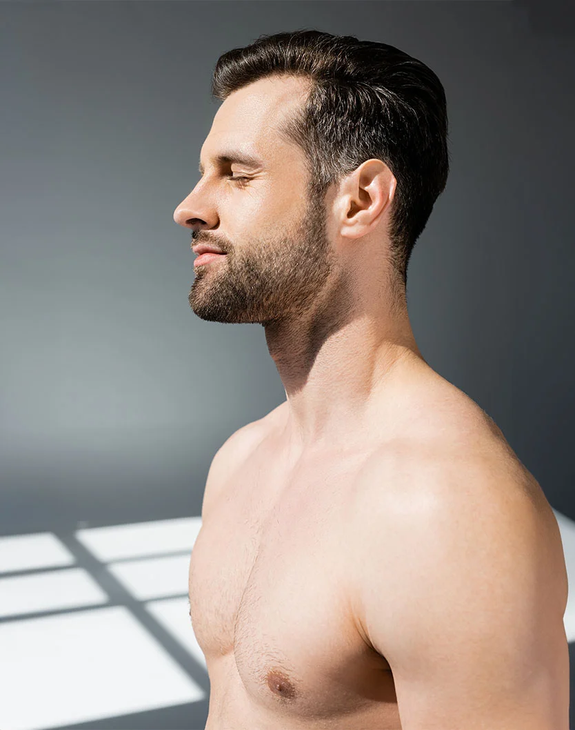 A profile view of a shirtless man with a light complexion, short dark hair, and a well-groomed beard. His eyes are closed, and his expression is calm. His defined shoulders and chest suggest a fit build, with light body hair visible. A neutral gray background highlights him, while soft lighting from above and to the side casts rectangular shadows on the floor, adding depth to the scene. - Gynecomastia in Toronto, Ontario