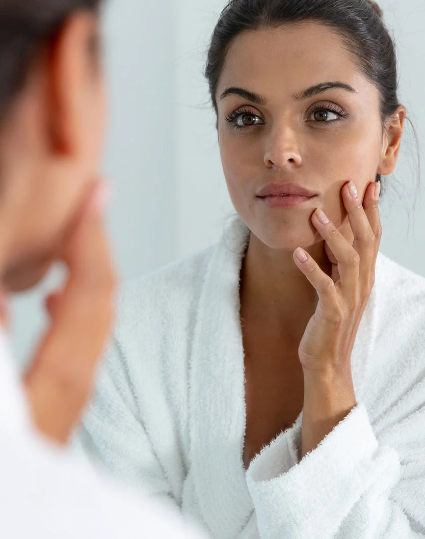A woman in a white robe gazes at her reflection in a mirror, gently touching her face. Her dark hair is pulled back in a bun, and her expression is introspective. A blurred reflection of another person is visible in the mirror. Soft, bright lighting highlights her smooth skin, creating a serene indoor setting. - Hydrafacial MD Facials in Toronto, Ontario
