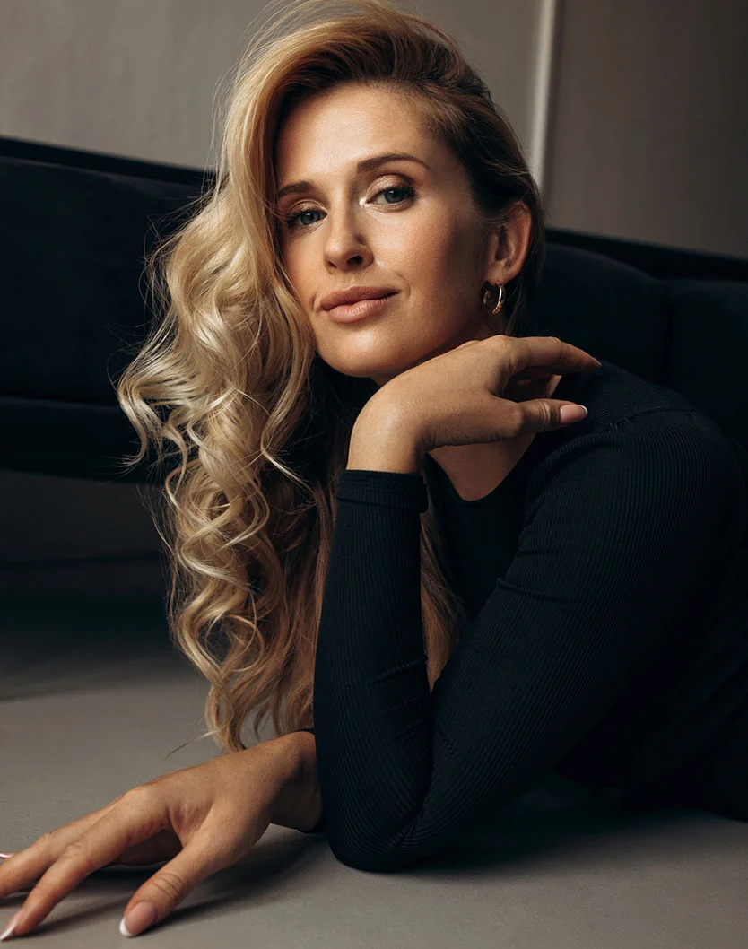 A woman with wavy blonde hair lies on a light gray surface, gazing at the viewer with a neutral expression. She wears gold hoop earrings and a black ribbed top. Soft, diffused lighting highlights her features and voluminous hair, creating an elegant, refined atmosphere against the muted gray background. - Hyperhidrosis in Toronto, Ontario