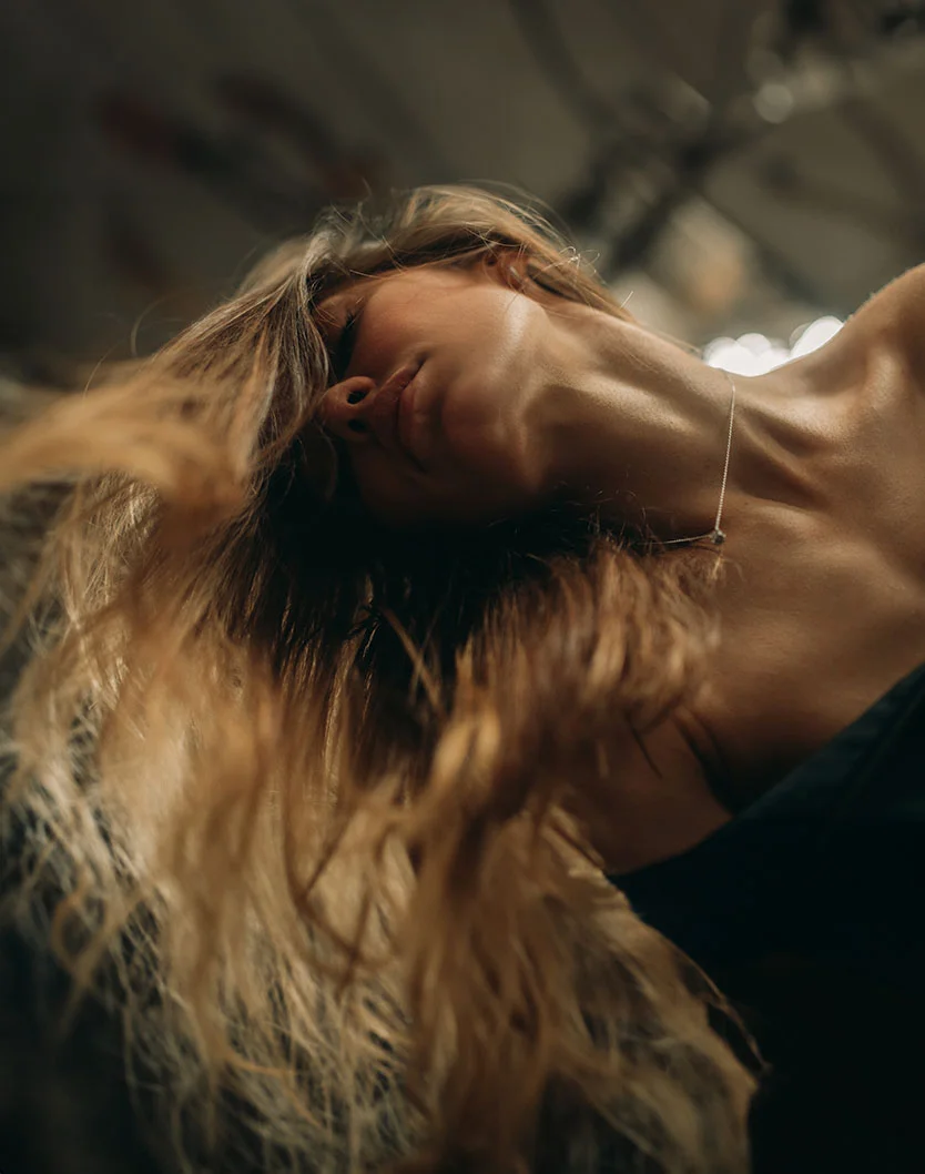 Close-up of a woman with long, light brown hair, slightly tousled and voluminous. She has a calm, contemplative expression, with her face angled downward. She wears a dark-colored top and a delicate necklace. The background is softly blurred, creating a warm, intimate atmosphere. - Jawline Filler in Toronto, Ontario