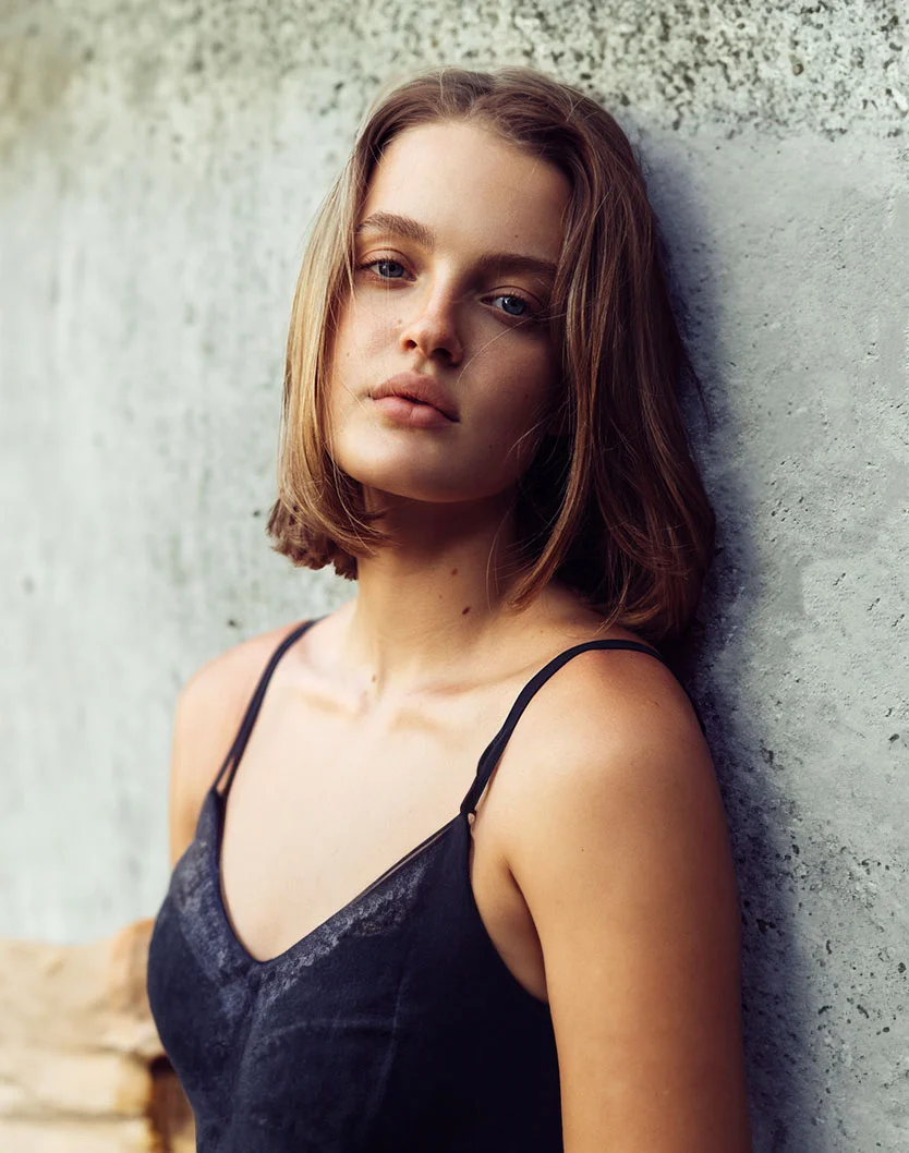 A young woman with shoulder-length brown hair stands against a light gray concrete wall. She gazes downward with a neutral expression, her light complexion highlighted by visible freckles. She wears a dark sleeveless top with lace detail on the bodice, which fits closely to her body. The textured concrete wall behind her has varied light gray tones. Soft, diffused lighting casts no harsh shadows, focusing on her face and upper body. - Lip Injections in Toronto, Ontario