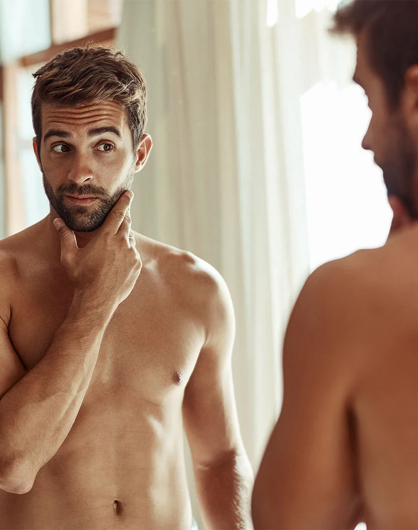 A light-skinned man with short, light brown hair and a beard stands shirtless in front of a mirror, looking thoughtfully at his reflection. Soft natural light highlights his toned body. The background features a window with sheer curtains, adding a warm ambiance. - Male Nipple Reduction in Toronto, Ontario