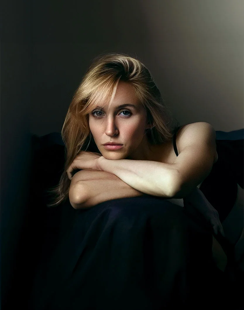 A close-up portrait of a woman with shoulder-length, light brown hair in soft waves. Her expression is contemplative, gazing slightly downward, with her lips closed and posture relaxed. She has fair skin, deep blue eyes, and well-defined eyebrows. She wears a dark top, blending with the dark background, which emphasizes her face. Soft, focused lighting highlights her features, creating a serene and subtle mood with gentle shadows. - Non-Surgical Face Lift in Toronto, Ontario