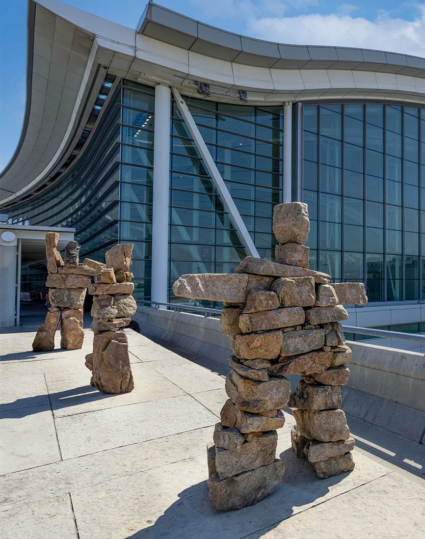 A full outdoor shot of a modern airport terminal features a light-gray structure with a curved, swooping roofline and large glass windows reflecting the bright blue sky. In front of the terminal, three large stone sculptures, resembling totem poles, are placed. The sculptures are made of stacked natural-colored rocks in shades of light brown, tan, and gray, representing human figures. The ground is covered in light-gray paving stones or concrete, with shadows cast by the sculptures and building under strong sunlight. - Out of Town in Toronto, Ontario