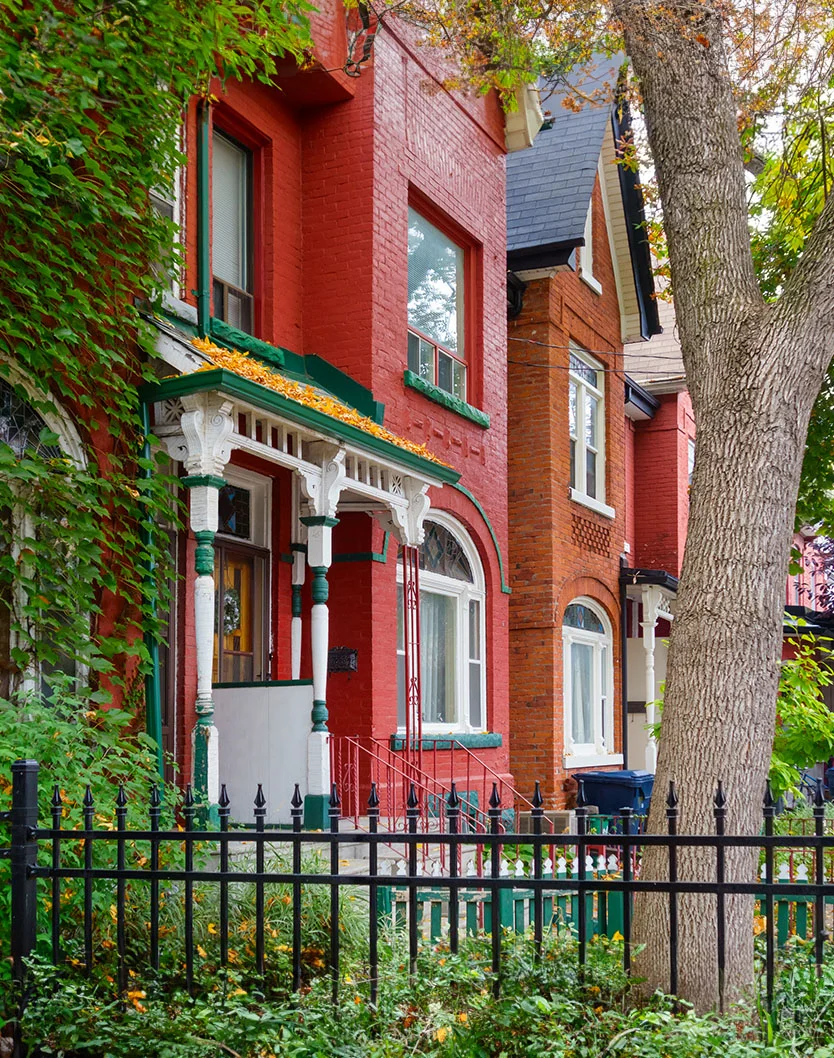 Two adjacent townhouses stand side by side. The left house is vibrant red brick with white trim and green-accented eaves, with vines climbing its side. The right house is a muted red-orange with white trim and a charcoal roof. A large tree with textured bark stands between them. A black metal fence with decorative finials lines the front, with greenery below. The lighting is natural, highlighting the houses and landscaping. - Out of Town Recovery in Toronto, Ontario