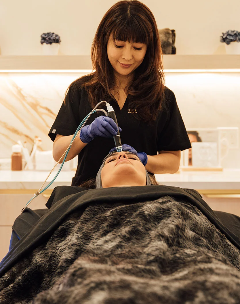 A woman, likely a medical professional, performs a facial treatment on a client lying on a table with a dark gray, textured blanket. Wearing a navy-blue top and blue gloves, she uses a skin treatment device connected to tubing. The client rests with closed eyes. The background is light beige with recessed shelves, light fixtures, and a small plant. - RF Microneedling in Toronto, Ontario