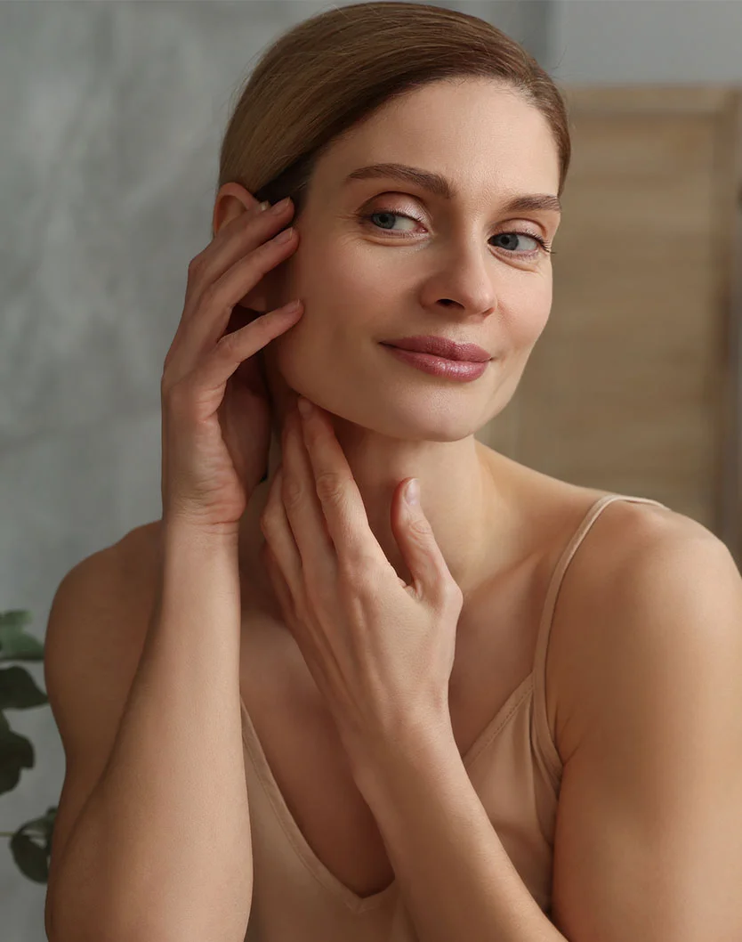 Close-up of a light-skinned woman with shoulder-length light brown hair, a neutral expression, and light-colored eyes. She touches her face and neck, wearing a beige tank top. The background is a soft grayish-beige with a hint of a leafy plant, lit by soft, diffused light. - Ultimate Signature Facial in Toronto, Ontario